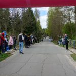 DSC_5630 Droga w lesie otoczona kibicami; biegacze na trasie. Wiosna, dominują zielenie i błękity. Nad drogą łuk startowy.