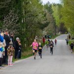 DSC_5633 Dzieci biegną w wyścigu ulicznym. Tło stanowi zielony las. Po lewej stronie stoi grupa widzów.