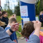 DSC_5636 Mężczyzna zakłada medal na szyję chłopca w różowej bluzie. W tle widoczni są ludzie i zielone drzewa. Scena jest na świeżym powietrzu.