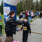 DSC_5640 Na zdjęciu jest bieg młodzieżowy na zewnątrz. Kobieta zakłada medal chłopcu w czarnym stroju z numerem 236. W tle są drzewa i kibice.