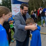 DSC_5643 Mężczyzna zakłada medal chłopcu w niebieskiej bluzie. W tle grupa ludzi na dworze, drzewa i plansza z mapą. Pogoda jest pochmurna.