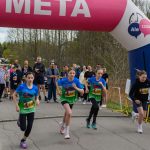 DSC_5650 Na zdjęciu grupa młodych biegaczy startuje spod różowo-białego łuku z napisem META. Tło stanowi zielony las i zebrani kibice. Atmosfera zawodów sportowych na ścieżce asfaltowej.