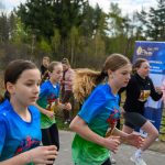 DSC_5654 Kilka dziewcząt biegnie w wyścigu, ubrane w niebieskie koszulki. W tle widoczne są drzewa i kibice. Obok stoi niebieski baner reklamowy.