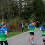 DSC_5655 Cztery osoby biegną wzdłuż asfaltowej drogi w lesie; mają kolorowe koszulki. Tło to zieleń drzew i krzewów. Kilka osób obserwuje bieg.