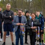 DSC_5658 Grupa ludzi stoi na tle drzew, trzymając medale. Są ubrani kolorowo. Tło jest zielone, jak w parku na wiosnę.
