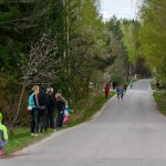 DSC_5661 Biegacze na asfaltowej drodze w otoczeniu zielonego lasu, obserwowani przez grupę osób. Po bokach drogi widoczne drzewa. Chmury na niebie.