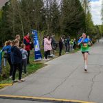 DSC_5663 Biegacz w zielonej koszulce z numerem 261 biegnie po asfaltowej drodze. W tle drzewa i kibice stoją pod niebieskim banerem. Impreza sportowa na świeżym powietrzu.