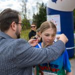 DSC_5668 Mężczyzna zakłada nastolatce medal na niebieskiej koszulce. Tło to wiejska droga z drzewami i niebiesko-białym dmuchanym łukiem. W oddali widać grupkę osób.