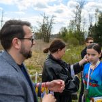 DSC_5669 Na zdjęciu widoczne jest dziecko w niebieskiej koszulce z medalem. W tle dorosłe osoby i zielone drzewa. Scena ma miejsce na świeżym powietrzu, z widocznymi chmurami na niebie.
