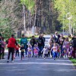 DSC_5670 Grupa dzieci biegnąca po asfaltowej drodze w otoczeniu lasu. W tle są drzewa i dorośli kibicujący uczestnikom. Dzieci mają kolorowe ubrania i numery startowe na koszulkach.
