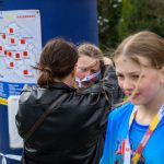 DSC_5672 Na zdjęciu są dzieci z medalami po zakończeniu biegu. Kolorowe stroje na tle zieleni i ludzie w tle. Na obok widoczna jest mapa z kalendarzem.