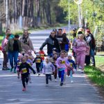 DSC_5675 Grupa małych dzieci uczestniczy w biegu ulicznym. W tle las, dominują kolory zieleni i błękitu. Dzieci mają numery startowe.