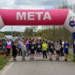 DSC_5679 Zdjęcie przedstawia start biegu z dziecięcymi uczestnikami pod różowo-niebieskim łukiem z napisem META. W tle widać drzewa i samochody, a z boku stoi mężczyzna z mikrofonem. Na zegarze widnieje czas 0:00:00