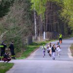 DSC_5680 Grupa dzieci biegnie po asfaltowej drodze otoczonej zielonym lasem. Na poboczu stoi kilku strażaków w czarnych mundurach. Tło tworzą drzewa z wiosennym listowiem.
