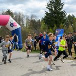 DSC_5687 Grupa dzieci rozpoczyna bieg przy różowo-niebieskim łuku startowym. Ubrane są w sportową odzież, niektóre w jaskrawe kolory. W tle widać drzewa i ludzi kibicujących.