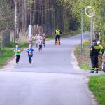 DSC_5688 Na zdjęciu grupa dzieci biegnie po asfaltowej drodze otoczonej zielonym lasem. W tle stoi dwóch strażaków i ochroniarz. Dzieci mają kolorowe koszulki i numery startowe.