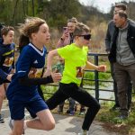 DSC_5689a Dzieci biegną w zawodach na zewnątrz. Tło to zielony krajobraz i widzowie. Ubrani są w sportowe stroje w żywych kolorach.