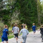DSC_5690 Dzieci biegają po ścieżce. Ubrane w kolorowe stroje sportowe, otoczone zielenią drzew. Droga prowadzi przez las.