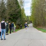 DSC_5694 Biegacz w niebieskim stroju biegnie po asfaltowej drodze w lesie. W tle widać drzewa i dopingujących ludzi. Po lewej stronie stoi niebieski baner.