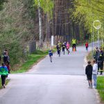 DSC_5699 Na drodze między drzewami biegnie grupa dzieci, ubranych w kolorowe stroje sportowe. Po bokach stoją dorośli, niektórzy w ciemnych ubraniach. Tło jest zdominowane przez zieleń i wysokie drzewa.