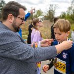 DSC_5700 Mężczyzna wręcza medal chłopcu w koszulce z numerem 259. W tle osoby, zielone krzewy i pochmurne niebo. Atmosfera sportowej uroczystości.