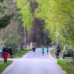 DSC_5702 Na zdjęciu widoczni są biegnący ludzie na leśnej drodze. Tło stanowią zielone drzewa i krzewy. Wokół znajdują się osoby obserwujące oraz zaparkowany samochód.
