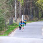 DSC_5708 Na zdjęciu widoczne są cztery osoby biegnące drogą, ubrane w niebiesko-zielone koszulki. Po lewej stronie znajduje się zalesiony teren z zielonymi drzewami. Tło stanowi las z wysokimi drzewami i ogrodzenie wzdłuż drogi.