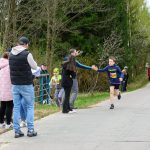 DSC_5709 Na zdjęciu młody biegacz w niebieskim stroju z numerem 264 biegnie po asfaltowej drodze. Tło stanowi zielony las i widzowie z boku. Obok drogi widoczny jest niebieski baner reklamowy.