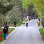 DSC_5715 Biegacze na asfaltowej drodze pośród zielonych drzew. W tle widać zarośla i strażaków w odblaskowych kamizelkach. Jasny dzień, spokojne otoczenie.