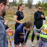 DSC_5716 Na zdjęciu grupa ludzi po biegach, dziecko w zielonej czapce podaje wodę. Tło to sucha roślinność, część osób nosi medale. Kolory są jasne, dominują zielenie i żółcie.