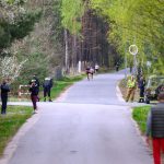 DSC_5719 Widać asfaltową drogę przez las, ludzie biegną i spacerują. Po bokach dużo zieleni i drzewa. Ochrona i samochód po prawej.