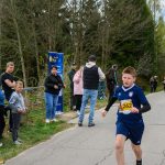 DSC_5719a Na zdjęciu chłopiec w niebieskim stroju biegnie po asfalcie. Obok stoi grupa kibiców przy ogrodzeniu. W tle widać drzewa i niebo.