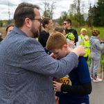 DSC_5720 Grupa ludzi na zewnątrz, część w sportowych ubraniach. Mężczyzna w marynarce przytula chłopca w koszulce z numerem. W tle drzewa i niebo.