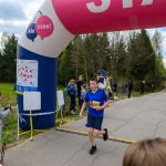 DSC_5725 Na zdjęciu widoczny jest biegacz w niebieskiej koszulce, przekraczający linię mety z numerem startowym 263. Tło stanowi zalesiona sceneria z widocznym niebem i publicznością. Nad linią mety wisi różowy i niebieski balon z nap