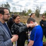 DSC_5727 Na zdjęciu kobieta zakłada medal chłopcu w niebieskiej koszulce. W tle ludzie na tle zieleni i lekko zachmurzone niebo. Atmosfera jest radosna i uroczysta.