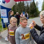 DSC_5737 Na zdjęciu dzieci na zawodach biegowych, przyjmujące medal od uśmiechniętej osoby. W tle nadmuchiwany łuk i zalesiona sceneria. Kolory są jasne i wiosenne, dominują zielony i niebieski.