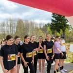 DSC_5740 Grupa młodych osób w czarnych strojach sportowych stoi pod kolorowym łukiem startowym. W tle drzewa i niebo zasłane chmurami. Na numerach startowych widoczne numery i logo sponsorów.