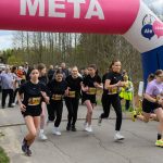 DSC_5749 Grupa młodych kobiet biegnie w zawodach pod różowo-białym balonem z napisem META. W tle las i kibice. Start wyścigu oznaczony zegarem.