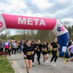 DSC_5750 Na zdjęciu widać grupę biegaczek przekraczających linię mety pod różowo-białym łukiem z napisem META. Tło stanowi zielony krajobraz z drzewami i tłumem widzów. Ubrania biegaczek są głównie czarne, z
