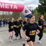 DSC_5752 Na zdjęciu młode biegaczki biegną przez linię mety. Nad nimi różowy łuk z napisem „META”. W tle widoczni są kibice i drzewa.