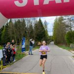 DSC_5764 Biegaczka przekracza linię mety pod różowym, nadmuchiwanym łukiem z napisem START. W tle drzewa i kibice. Dzień jest pochmurny.