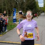 DSC_5765 Biegaczka w fioletowej koszulce z numerem 269 biegnie drogą. Ma wielokolorowe okulary. W tle las i kibicująca grupa ludzi.