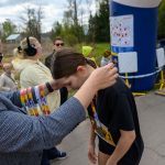 DSC_5770 Mężczyzna wręcza medal dziewczynie na mecie biegu. W tle drzewa i balon reklamowy. Kilka osób obserwuje z boku.