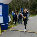 DSC_5783 Na obrazku kobieta w czarnym stroju sportowym biegnie przez linię mety. W tle widać kibiców i zielone drzewa. Po lewej stronie znajduje się niebieski balon startowy z mapą.