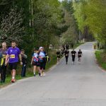 DSC_5788 Grupa biegaczy na asfaltowej drodze w lesie. Ubrani w kolorowe stroje z numerami startowymi. Tło zielone, otoczone drzewami.
