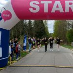 DSC_5790 Początek biegu pod różowym balonem z napisem START. Biegacze w czarnych strojach trzymają się za ręce. W tle drzewa i kibice na poboczu.