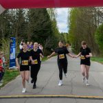 DSC_5795 Na zdjęciu cztery biegaczki przekraczają linię mety, trzymając się za ręce. Ubrane na czarno, na tle zielonych drzew. Dookoła widzowie i niebiesko-różowy baner.