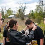 DSC_5804 Na zdjęciu osoba zakłada medal na szyję uczestniczki biegu. W tle zielone krzewy i dom przy pochmurnym niebie. Ubrani są w czarne koszulki.