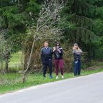 DSC_5807 Trzy osoby stoją obok drogi w lesie, na tle zielonych drzew. Jedna z nich trzyma telefon w rękach. Scena jest wiosenna.
