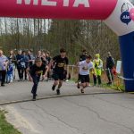 DSC_5809 Trzech młodych biegaczy startuje spod różowo-niebieskiego, dmuchanego łuku z napisem META. W tle widoczny jest tłum ludzi stojący na poboczu wśród drzew. Obok ustawiony jest elektroniczny zegar pomiarowy, wyświetlający czas